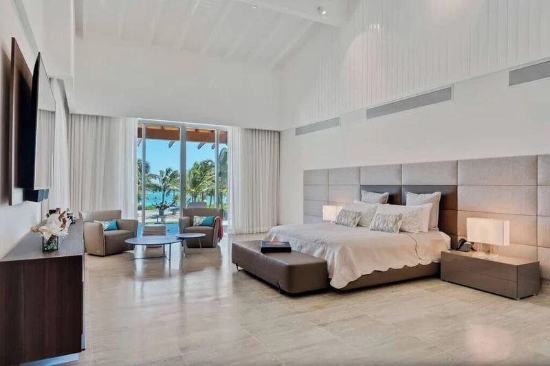 A modern hotel room with a large bed, a seating area with a coffee table, and a view of palm trees through the windows.