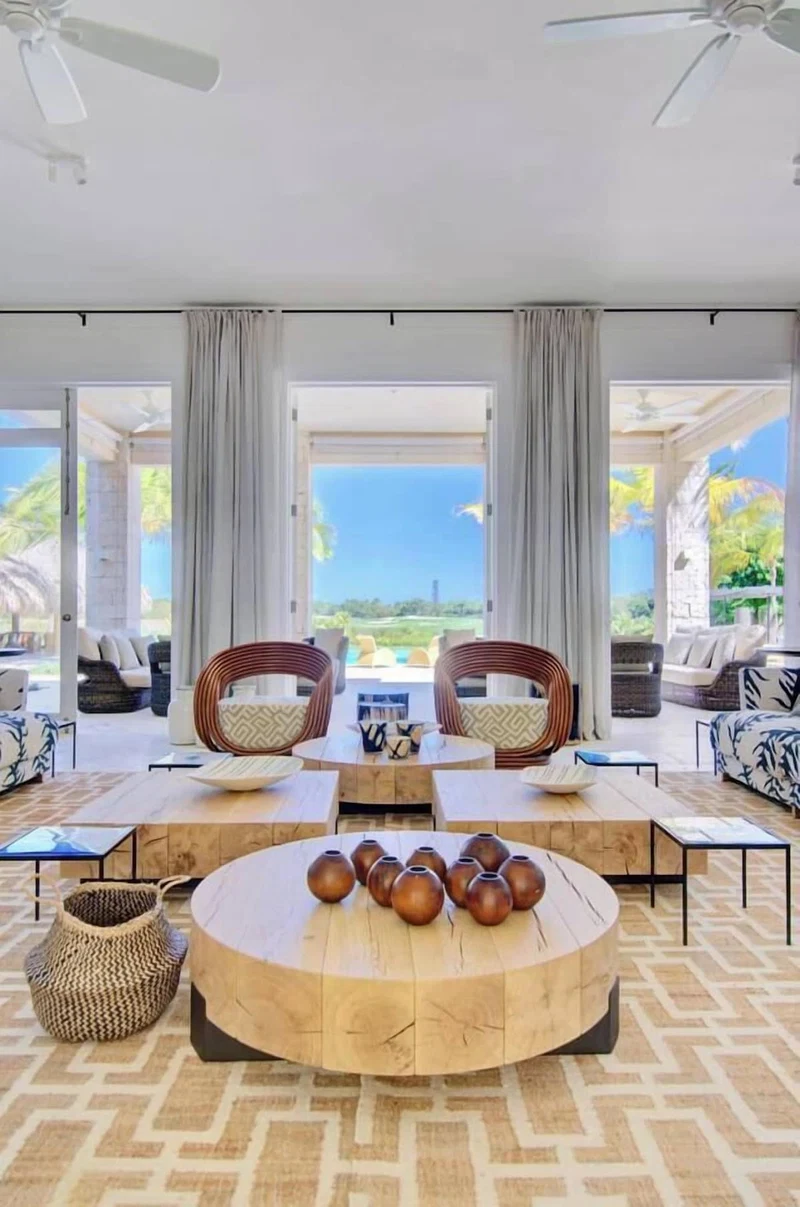 A modern living room with a round wooden coffee table, two wicker chairs, and a view of the ocean.