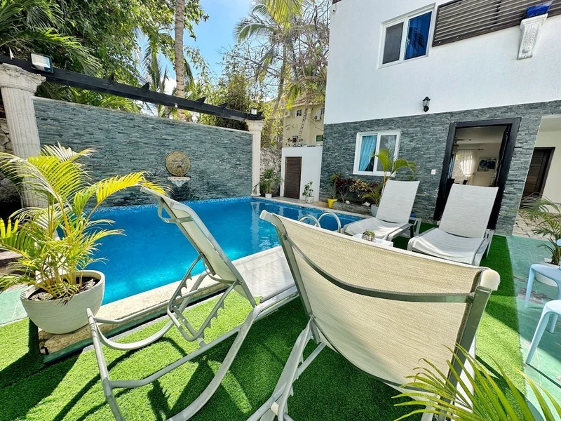A swimming pool with lounge chairs and artificial grass, surrounded by a stone wall and potted plants.