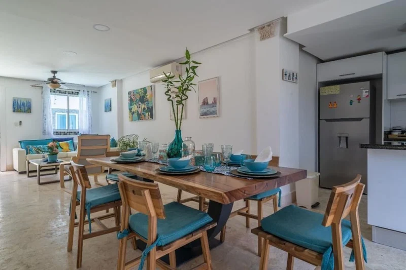 A dining area with a wooden table set for six, surrounded by chairs with teal cushions.