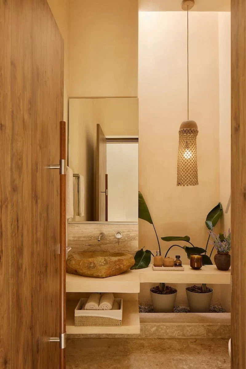 A bathroom with a wooden door, a round sink, a mirror, a hanging light fixture, and potted plants.