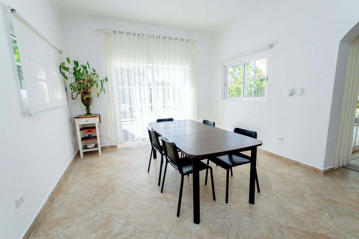 A room with a dining table, chairs, a potted plant, a whiteboard, and a window with curtains.