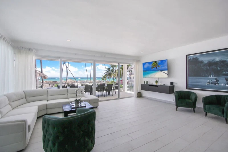 A modern living room with a white sectional sofa, a green ottoman, and a TV mounted on the wall. There is a large window with a view of the beach and palm trees.