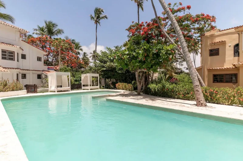 A pool area with a clear blue pool, surrounded by palm trees and red flowering plants.