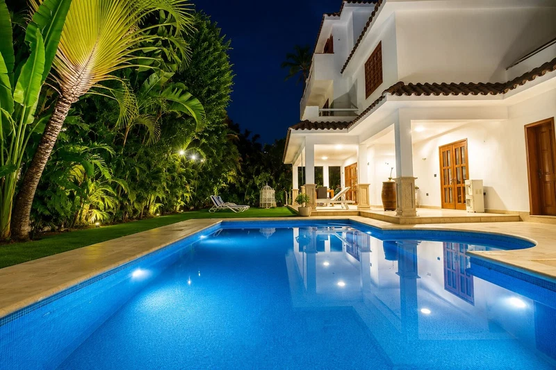 A swimming pool with a white house in the background, surrounded by greenery.