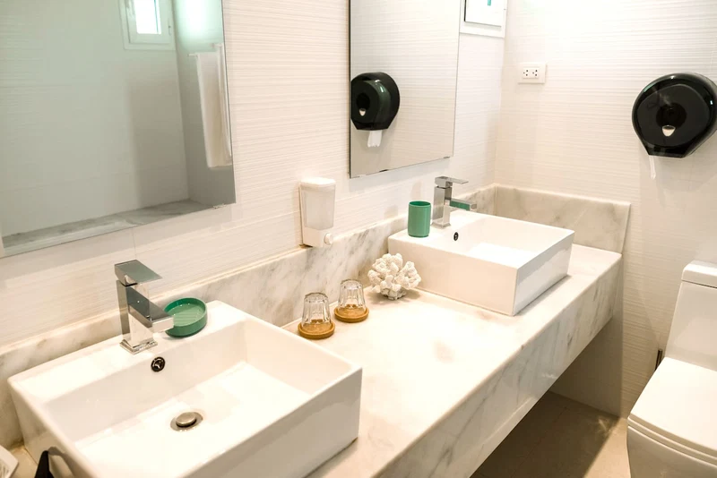 A bathroom with two sinks, a mirror, a toilet, and a soap dispenser.