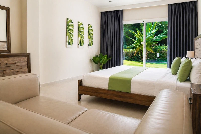 A bedroom with a bed, a sofa, a plant, and a window with a view of greenery.