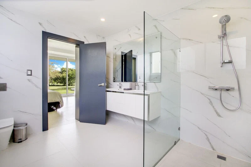 A modern bathroom with a glass shower enclosure, a white marble floor, and a view of a garden through an open door.