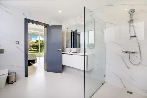 A modern bathroom with a glass shower enclosure, a white marble floor, and a view of a garden through an open door.