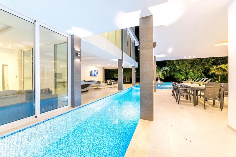A modern indoor pool area with glass walls, a dining area with outdoor seating, and a TV screen in the background.