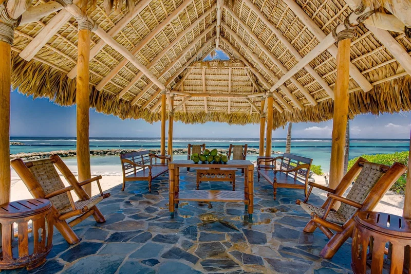 A beachside gazebo with a thatched roof, stone flooring, and wooden furniture.