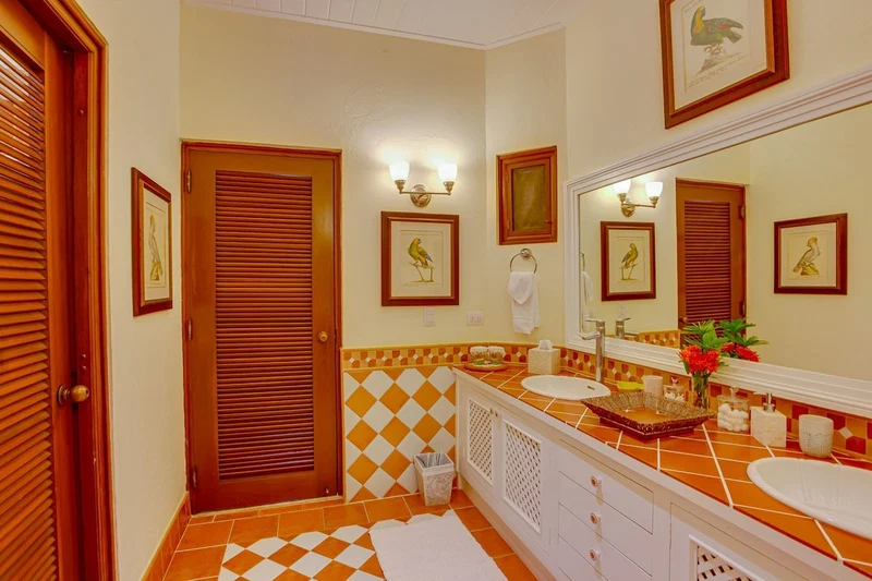 A bathroom with a wooden door, a tiled floor, a sink, a mirror, and framed pictures on the wall.