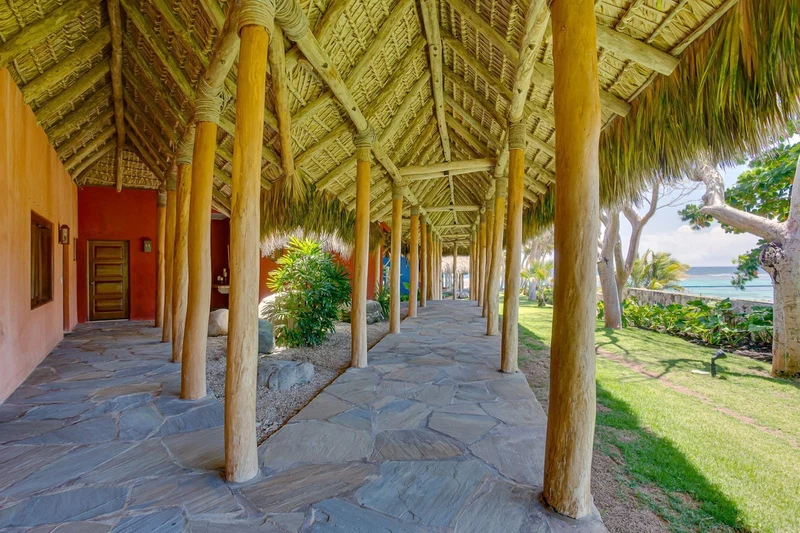 A covered walkway with stone flooring, wooden pillars, and a thatched roof.