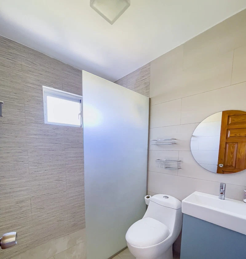 A bathroom with a modern design, featuring a toilet, a sink, and a mirror. There is a window providing natural light and a shower area with a glass door.