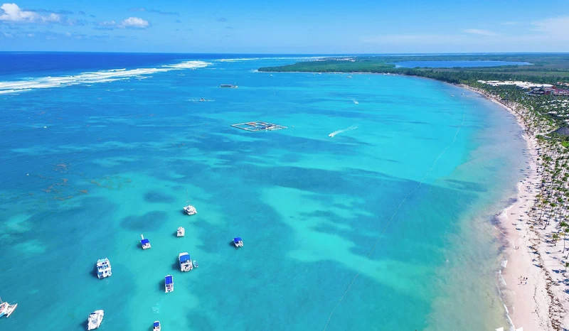 An aerial view of a coastal area with clear turquoise waters, a sandy beach, and several boats anchored near the shore.