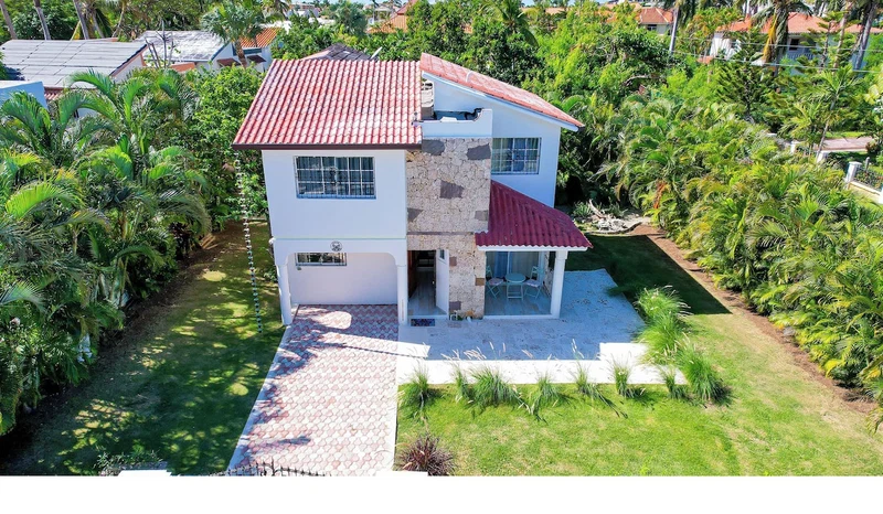 A two-story house with a red-tiled roof, surrounded by lush greenery and palm trees.