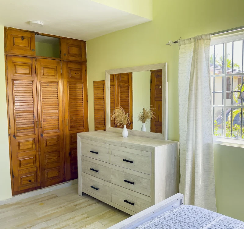 A bedroom with a wooden wardrobe, a dresser, a mirror, and a window with curtains.