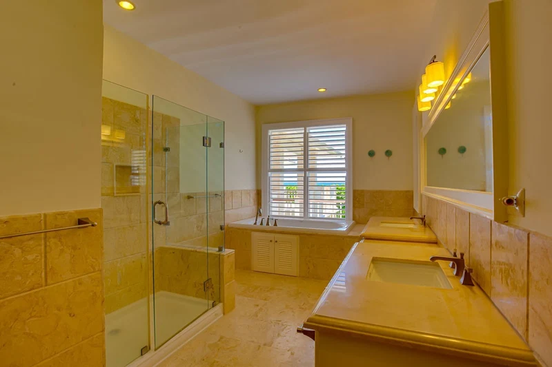 A modern bathroom with a glass-enclosed shower, a large window with blinds, and two sinks with mirrors.