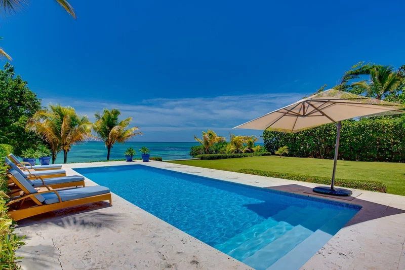 A swimming pool with lounge chairs and an umbrella, surrounded by greenery and a view of the ocean.
