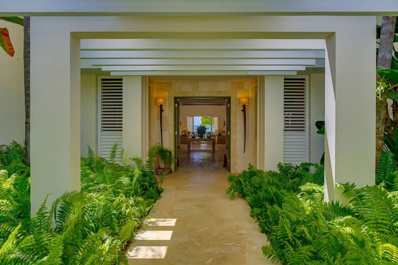 A pathway leading to a dining area with a view of the outdoors, flanked by lush greenery and white architectural elements.