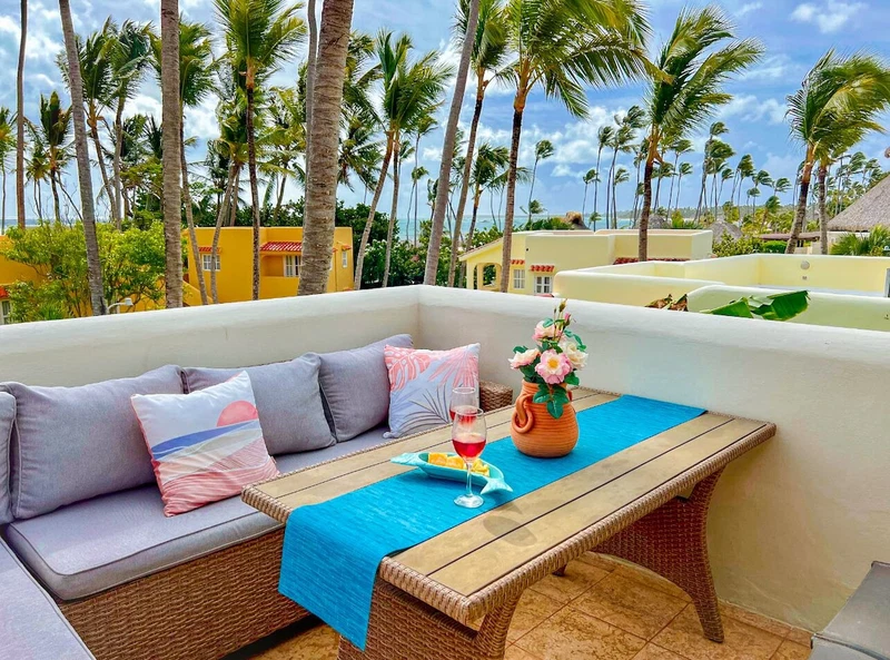 A sunlit outdoor seating area with a wicker sofa, a wooden table, and a vase of flowers.