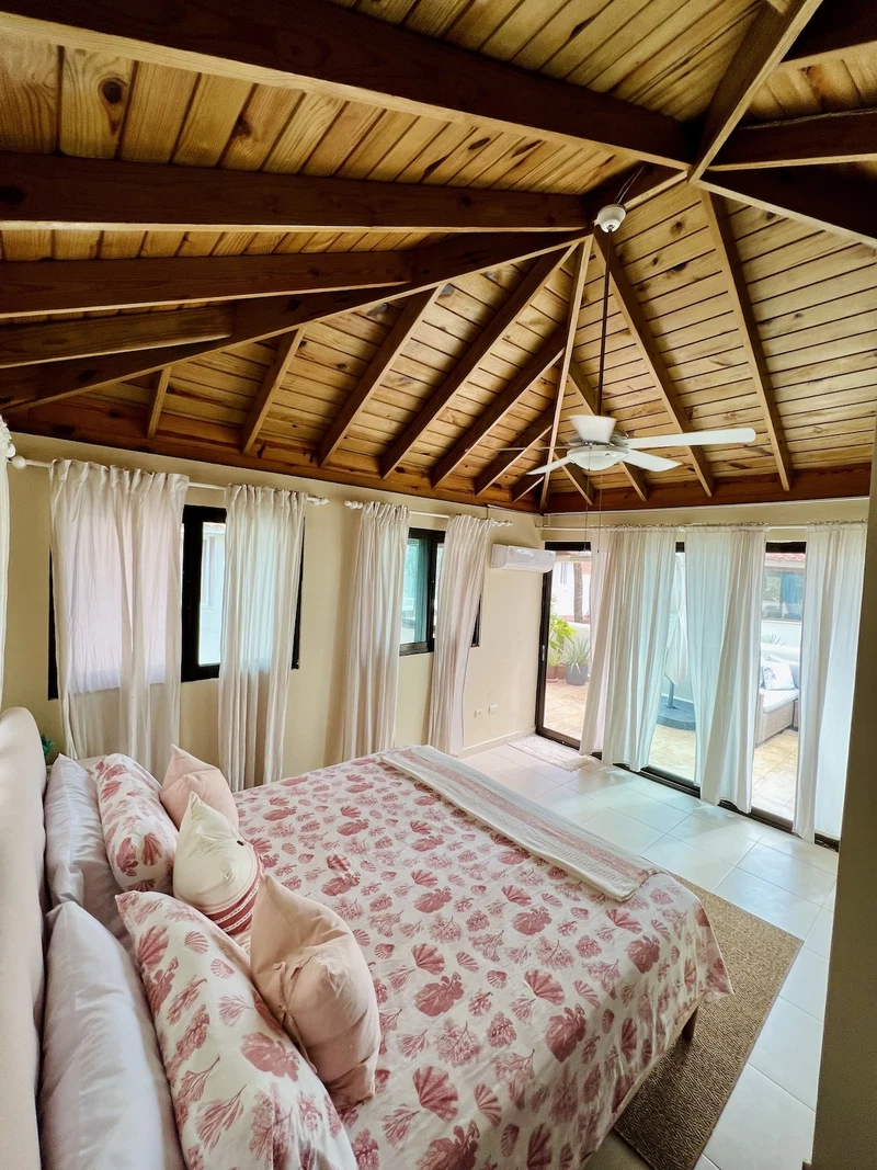 A bedroom with a wooden ceiling, a bed with floral bedding, and large windows with curtains.