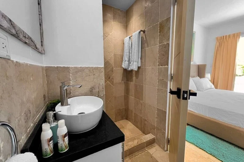 A hotel bathroom with a white sink, a shower, and a bed visible through an open door.