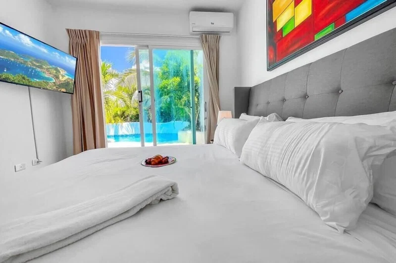 A hotel room with a bed, a flat-screen TV, a painting, and a view of a pool and greenery.