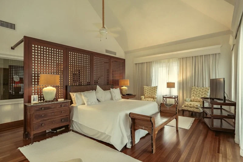 A spacious bedroom with a large bed, wooden headboard, two bedside tables with lamps, a wooden bench, a TV, and a large window with curtains.