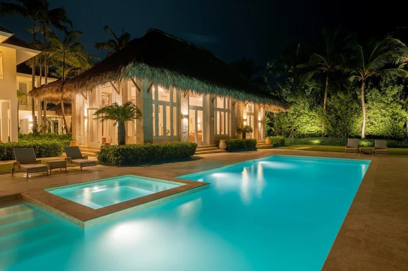 A poolside area with a clear blue pool, a thatched-roof structure, and palm trees.